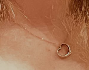 close-up photograph of a delicate, thin gold chain necklace resting against skin. The necklace features a small, open heart-shaped pendant that has a distinctive two-toned design, with one side in yellow gold and the other in white gold or silver.