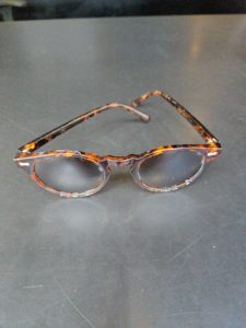 A pair of round, tortoiseshell-patterned glasses with a dark brown and amber mottled design, resting on a dark, textured grey surface. The frames feature small silver decorative pins at the hinges.