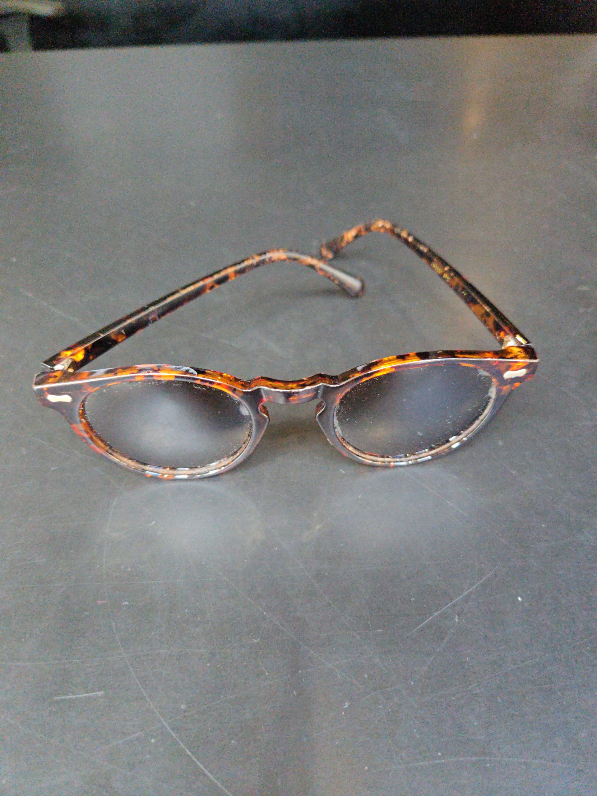 A pair of round, tortoiseshell-patterned glasses with a dark brown and amber mottled design, resting on a dark, textured grey surface. The frames feature small silver decorative pins at the hinges.
