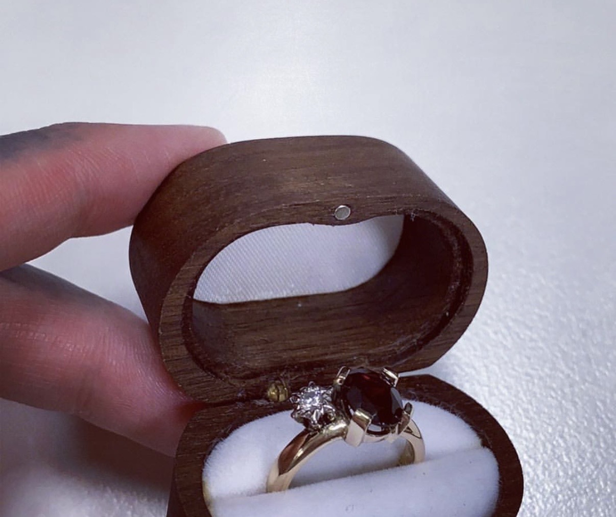 A gold ring with a large oval-cut dark red garnet in a claw setting next to a small clear diamond, presented inside an open wooden ring box.