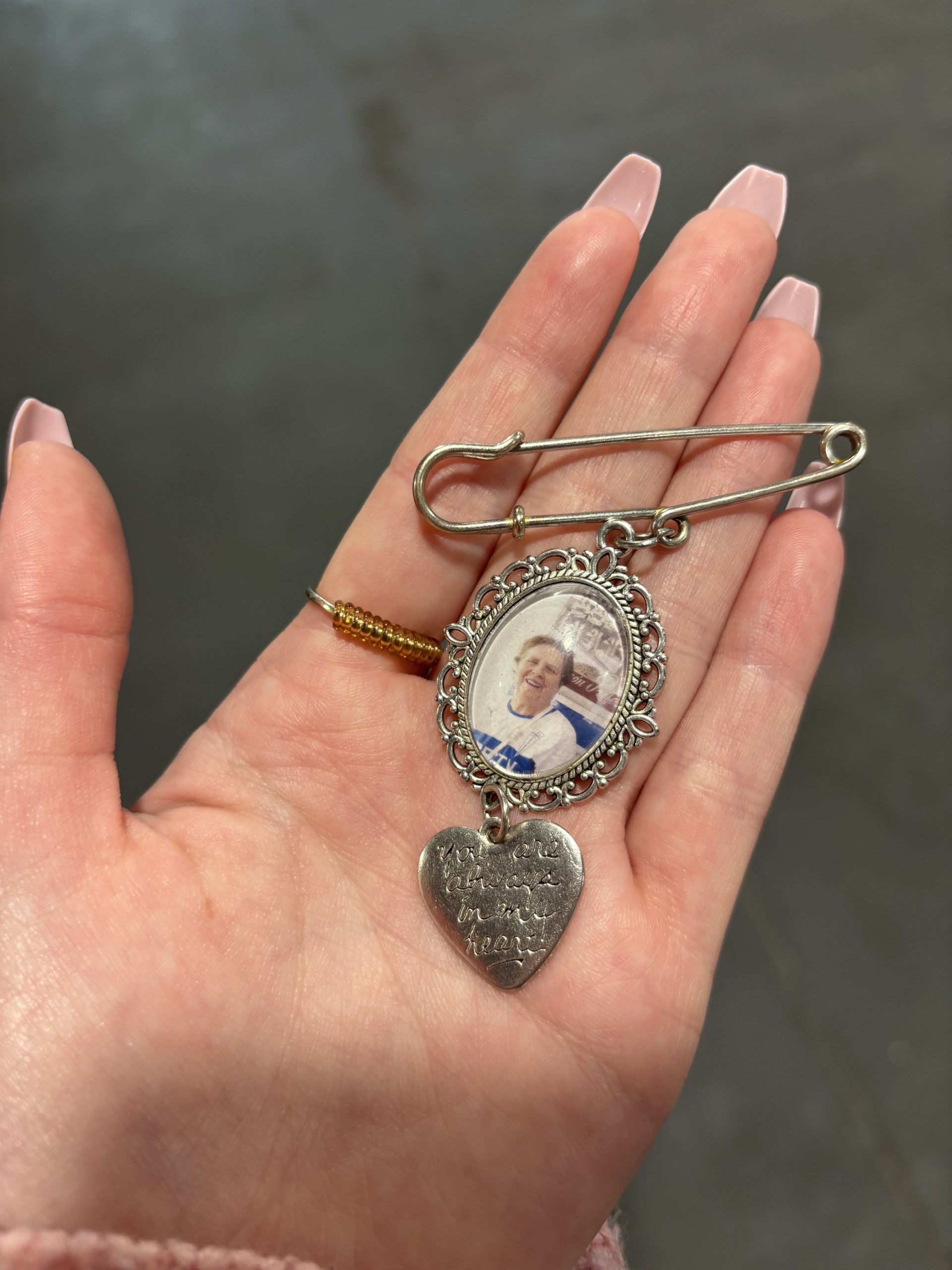 A silver safety-pin style memory brooch being held in a palm. The brooch features an oval, vintage-style filigree frame enclosing a photograph of a woman, with a hanging silver heart-shaped charm below it engraved with the text 'You are always in my heart'.